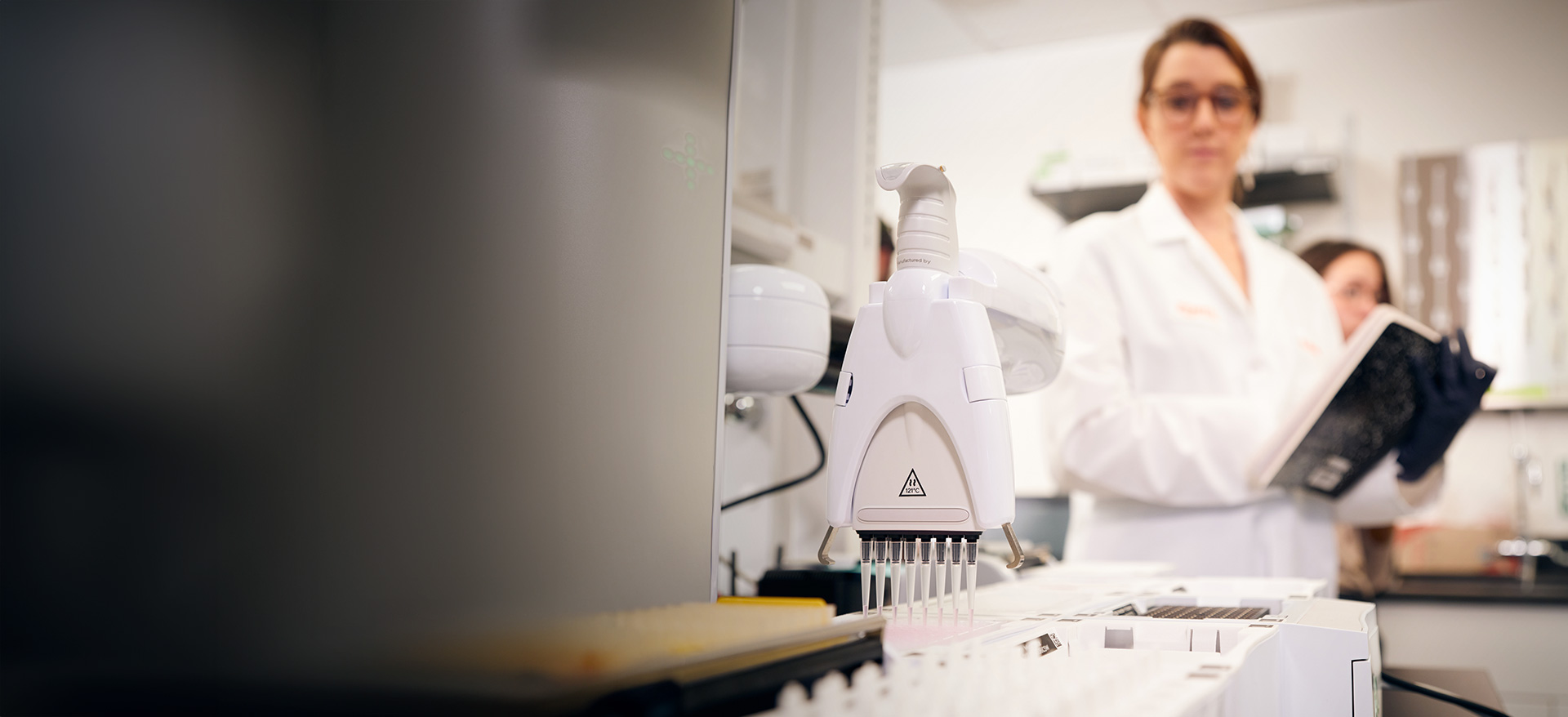 Close-up of an automated lab pipetting instrument dispensing into tubes, with a Zoetis researcher in a lab coat blurred in the background holding a clipboard - Zoetis