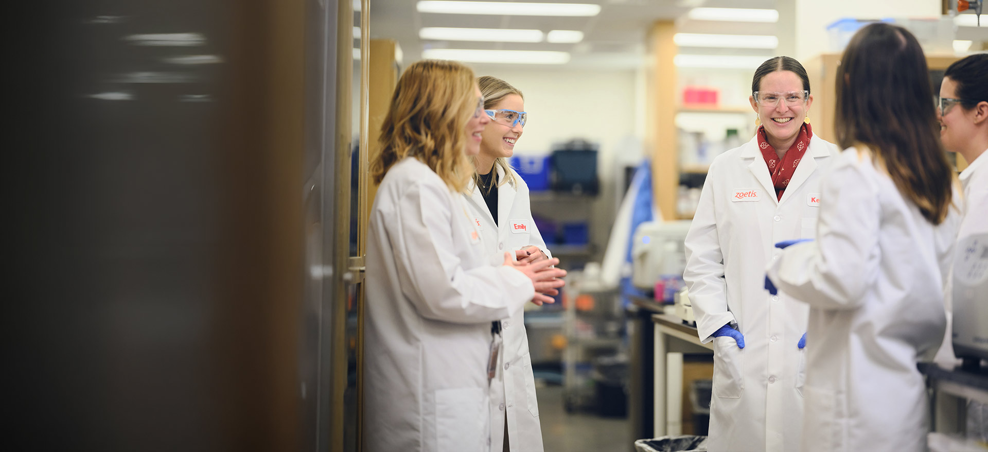 Group of Zoetis scientists in white lab coats and safety glasses chatting and smiling in a bright laboratory corridor - Zoetis